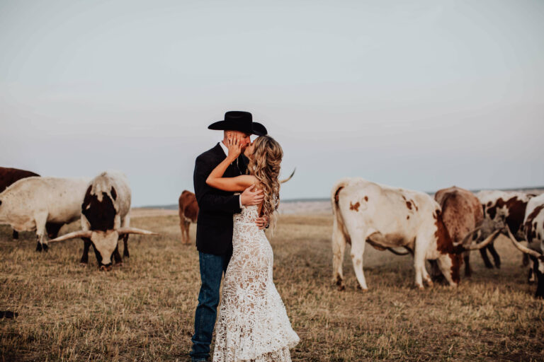 A bride and groom in a cowboy hat kiss in the pasture surrounded by Texas Longhorns at Younger Ranch