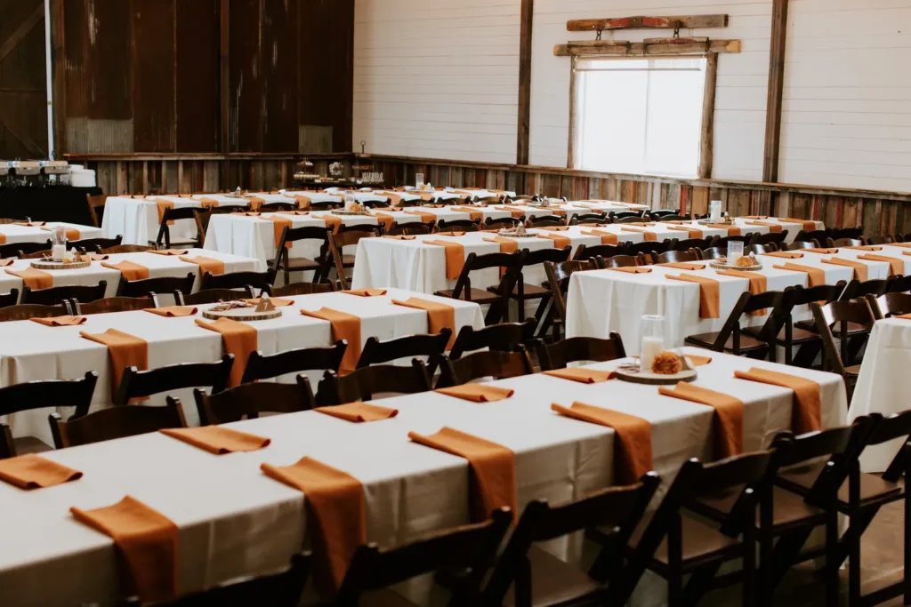 Long dinner tables with white linens, rust-orange napkins and wooden chairs set for a wedding reception inside the Reception Barn at Younger Ranch