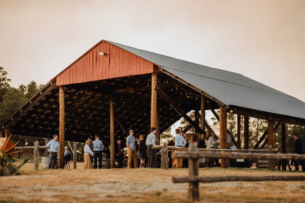 Wedding guests gathered for cocktail hour under the open-air hay barn at Younger Ranch, with string lights and a split-rail fence